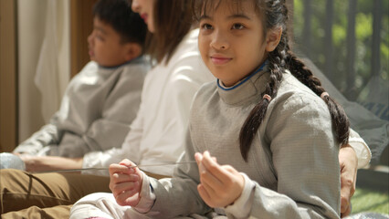 A girl and boy in fencing gear with their mother preparing food and sports equipment