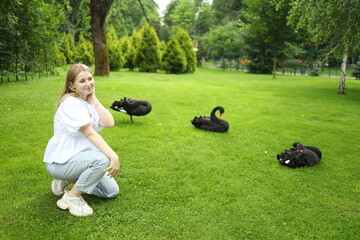 A young woman kneels on the grass in a vibrant park, playfully interacting with several black cats lounging around her. The sun shines through the trees, creating a serene atmosphere.
