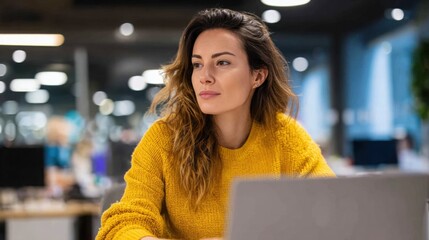 Medium shot of a woman in a casual yellow top looking attentively at her laptop screen, her hands on the keyboard, in a contemporary open-plan office setting.
