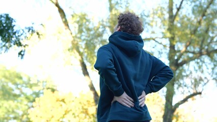 Back view Young Caucasian man massaging his back in green park between trees. Rear view sportsman...