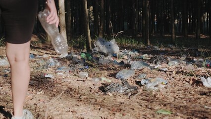 Woman walking through a pile of garbage in the forest and throwing a bottle