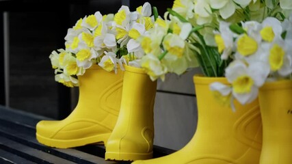 Bright yellow rubber boots creatively repurposed as vibrant springtime planters, overflowing with artificial white and yellow daffodil flowers on a slatted bench.
