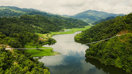 A stunning aerial view of a calm river winding through lush green hills, with a suspension bridge connecting both sides in a peaceful mountain valley.