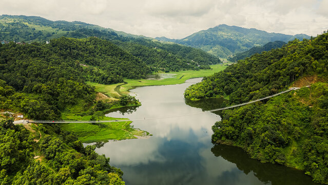 A stunning aerial view of a calm river winding through lush green hills, with a suspension bridge connecting both sides in a peaceful mountain valley.