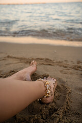 Sandy Feet with Anklet by the Sea. High quality photo. A close up of a woman's feet resting on the sandy beach, adorned with a stylish gold anklet featuring seashell and pearl charms. Gentle waves