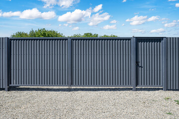 A modern gray corrugated metal fence with a built-in pedestrian gate stands on a gravel surface...