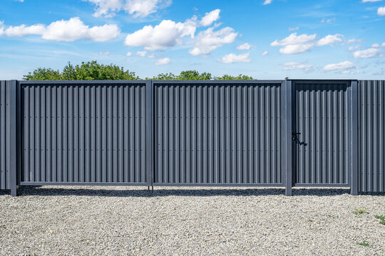 A modern gray corrugated metal fence with a built-in pedestrian gate stands on a gravel surface under a bright blue sky with scattered clouds.