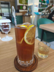 A tall glass of iced lemon tea with lemon slices and a stirrer, sits on a wooden coaster on a wooden table in a Surabaya cafe.