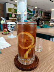 A tall glass of iced lemon tea with lemon slices and a stirrer, sits on a wooden coaster on a wooden table in a Surabaya cafe.