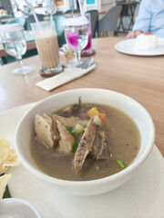 A white plate holds a bowl of Sop Buntut or Indonesian oxtail soup, with a side of chili paste, lime, and crispy emping crackers on a wooden table.
