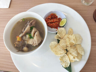 A white plate holds a bowl of Sop Buntut or Indonesian oxtail soup, with a side of chili paste, lime, and crispy emping crackers on a wooden table.