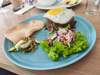A blue plate holds Indonesian fried rice with a sunny-side-up egg, a large prawn cracker, pickled vegetables, and fresh salad on a wooden table.