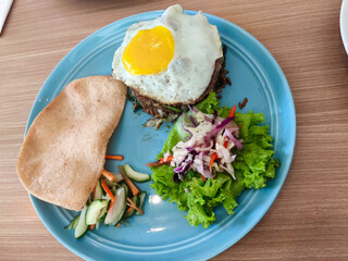 A blue plate holds Indonesian fried rice with a sunny-side-up egg, a large prawn cracker, pickled vegetables, and fresh salad on a wooden table.