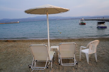 Relaxing beachside oasis with sun chairs, an umbrella, and boats gently bobbing in the water during a serene evening by the shore