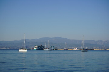 Serene sailboats glide across tranquil waters with a backdrop of distant mountains under a clear blue sky in the early evening light