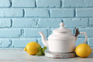 A serene still life featuring a pristine white teapot with a charming black handle, accompanied by two vibrant yellow lemons and fresh green leaves, set against the rustic texture of a blue brick wall