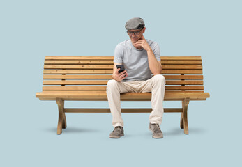 Obraz premium Smiling mature man in his 50s sitting on a park wooden bench and using a smartphone. Studio shot, isolated on blue background. Casual clothing, wearing reading glasses and a hat