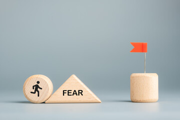 A wooden figure runs past a triangle block labeled FEAR toward a red flag, oncept of overcoming fear and challenges to achieve goals and success