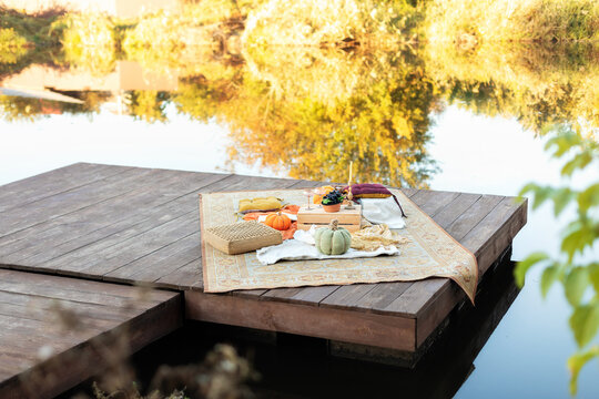 Aesthetic boho autumn picnic on the Pierce with Lake with pumpkins, carpet, plaid, cushions, fall decor, book, fruits, table with glasses. Cozy outdoors spase for relax near river. Party decoration	
