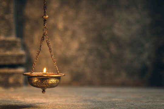 A glowing brass diya hanging from an iron chain, suspended in mid-air against a dim wall, casting a soft golden shadow on the background, traditional temple-style decor