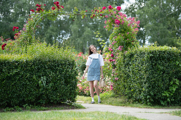 A young woman in a denim dress strolls along a path in a lush rose garden on a sunny day,...