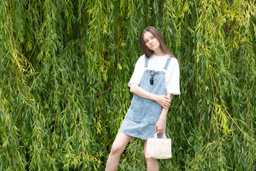 Young model stands confidently against a vibrant green foliage backdrop, wearing a denim dress and...
