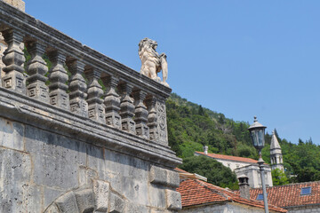 Sunny Perast street scene with traditional stone buildings, a charming cafe with outdoor seating, and lush greenery.