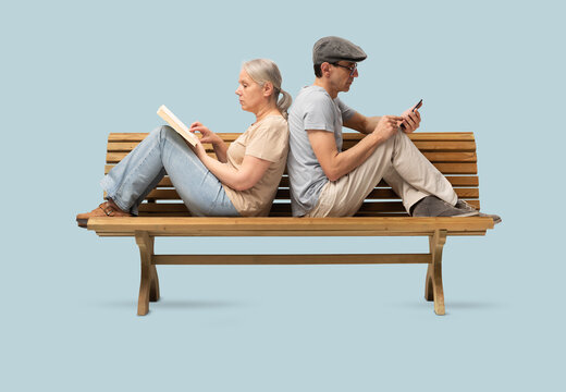 Mature couple sitting back to back on a wooden bench, woman reading book and man using smartphone. Concept of  emotional distance in modern relationships. Studio shot isolated on blue background.