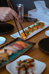 Hand Picking Up Fried Shrimp with Chopsticks. A hand uses chopsticks to pick up a crispy fried shrimp from a white rectangular plate. Other plates of sushi and food are visible on the wooden table.