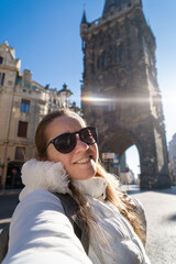 Mujer turista paseando por la ciudad de Praga y tomándose una selfie, con la torre de la pólvora y el sol naciente de fondo.	