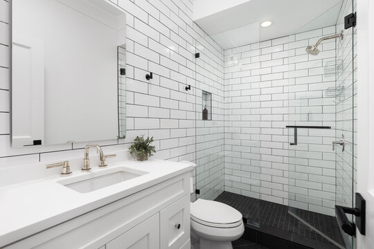 A white bathroom with subway tile walls, gold faucets, and black square tile flooring. - Powered by Adobe