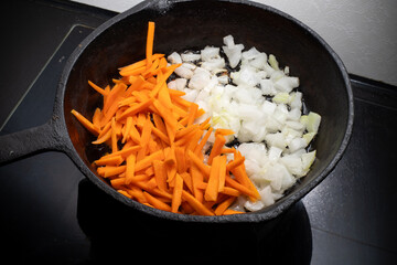 Home cooking. On the stove in a cast iron frying pan onions and carrots are fried in a hot frying...