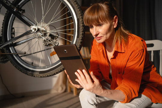 Middle aged woman repairing a broken bicycle at home. Female cyclist inspecting bicycle with digital tablet.