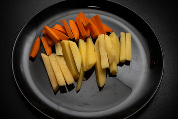 Raw potatoes cut into large strips and raw carrots cut into strips on a black plate, top view, side...