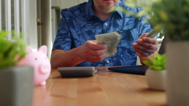 Man Counts His 100 Dollar Bills Methodically, Surrounded By A Familiar Kitchen Setting That Reflects Real-Life Financial Routines And Daytime Stillness.Male Hands Managing Cash On Kitchen Table 