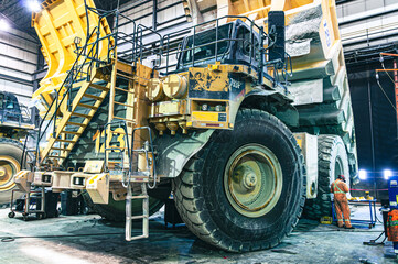 Massive mining haul truck undergoing maintenance in an industrial garage. A worker in an orange suit provides scale to this powerful piece of heavy machinery.
