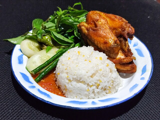 Lalapan ayam goreng. Crispy golden fried chicken dishes, with bright red chili sauce, fresh cucumber slices and a few basil leaves on a white enamel plate decorated with blue.