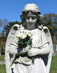 A serene stone angel holding white roses in a sunlit cemetery