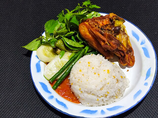 Lalapan ayam goreng. Crispy golden fried chicken dishes, with bright red chili sauce, fresh cucumber slices and a few basil leaves on a white enamel plate decorated with blue.