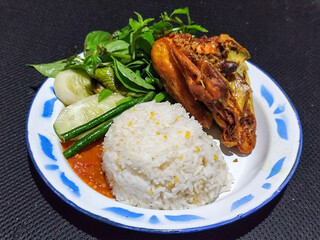 Lalapan ayam goreng. Crispy golden fried chicken dishes, with bright red chili sauce, fresh cucumber slices and a few basil leaves on a white enamel plate decorated with blue.