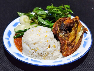 Lalapan ayam goreng. Crispy golden fried chicken dishes, with bright red chili sauce, fresh cucumber slices and a few basil leaves on a white enamel plate decorated with blue.