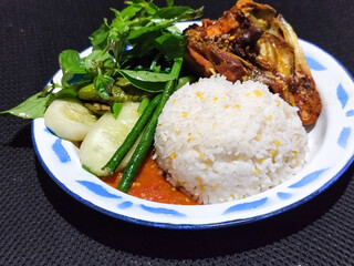 Lalapan ayam goreng. Crispy golden fried chicken dishes, with bright red chili sauce, fresh cucumber slices and a few basil leaves on a white enamel plate decorated with blue.