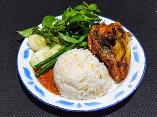 Lalapan ayam goreng. Crispy golden fried chicken dishes, with bright red chili sauce, fresh cucumber slices and a few basil leaves on a white enamel plate decorated with blue.