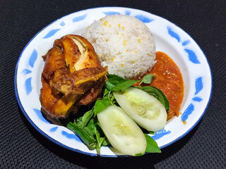 Lalapan ayam goreng. Crispy golden fried chicken dishes, with bright red chili sauce, fresh cucumber slices and a few basil leaves on a white enamel plate decorated with blue.