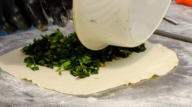 A baker uses a spoon to place finely chopped green herb filling from a bowl onto a layer of rolled dough Preparing vegan pastry Vegetarian flatbread Jingyalovhats traditional Armenian food