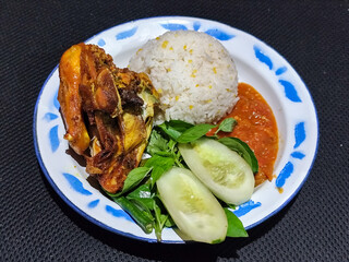 Lalapan ayam goreng. Crispy golden fried chicken dishes, with bright red chili sauce, fresh cucumber slices and a few basil leaves on a white enamel plate decorated with blue.
