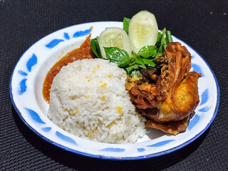 Lalapan ayam goreng. Crispy golden fried chicken dishes, with bright red chili sauce, fresh cucumber slices and a few basil leaves on a white enamel plate decorated with blue.