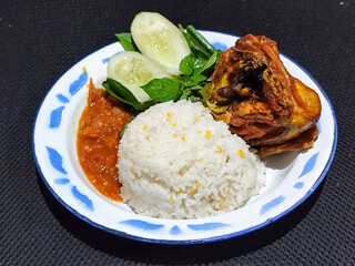 Lalapan ayam goreng. Crispy golden fried chicken dishes, with bright red chili sauce, fresh cucumber slices and a few basil leaves on a white enamel plate decorated with blue.