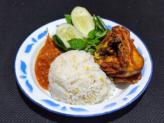Lalapan ayam goreng. Crispy golden fried chicken dishes, with bright red chili sauce, fresh cucumber slices and a few basil leaves on a white enamel plate decorated with blue.