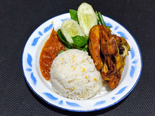 Lalapan ayam goreng. Crispy golden fried chicken dishes, with bright red chili sauce, fresh cucumber slices and a few basil leaves on a white enamel plate decorated with blue.